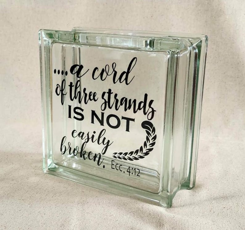 May include: A square, clear glass block featuring black lettering that reads, "...a cord of three strands IS NOT easily broken. Ecc. 4:12," accompanied by a decorative leaf-like flourish at the end of the text, creating a transparent display piece.