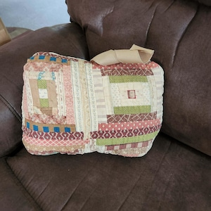 May include: A rectangular throw pillow featuring a patchwork design made of various fabric strips in shades of pink, green, blue, and cream. The surface displays multiple patterns including floral, striped, and checkered prints, topped with a beige ribbon.