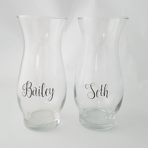 May include: Two clear glass hurricane vases with a flared rim and a rounded base. Each vase features black cursive lettering on the front, with one vase displaying the name Bailey and the other vase displaying the name Seth in a matching font.