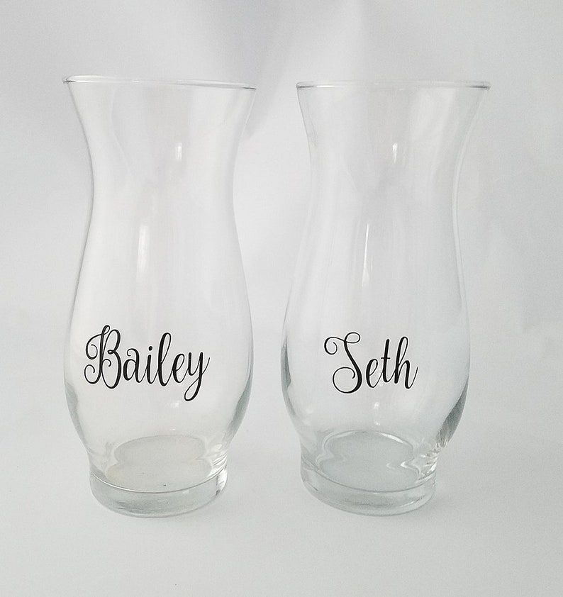 May include: Two clear glass hurricane vases with a flared rim and a rounded base. Each vase features black cursive lettering on the front, with one vase displaying the name Bailey and the other vase displaying the name Seth in a matching font.