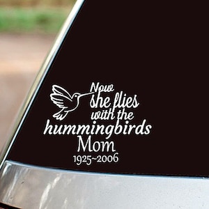 May include: White vinyl decal featuring a hummingbird and the text "Now she flies with the hummingbirds Mom 1925-2006". The decal is designed for application on a car window or similar surface.