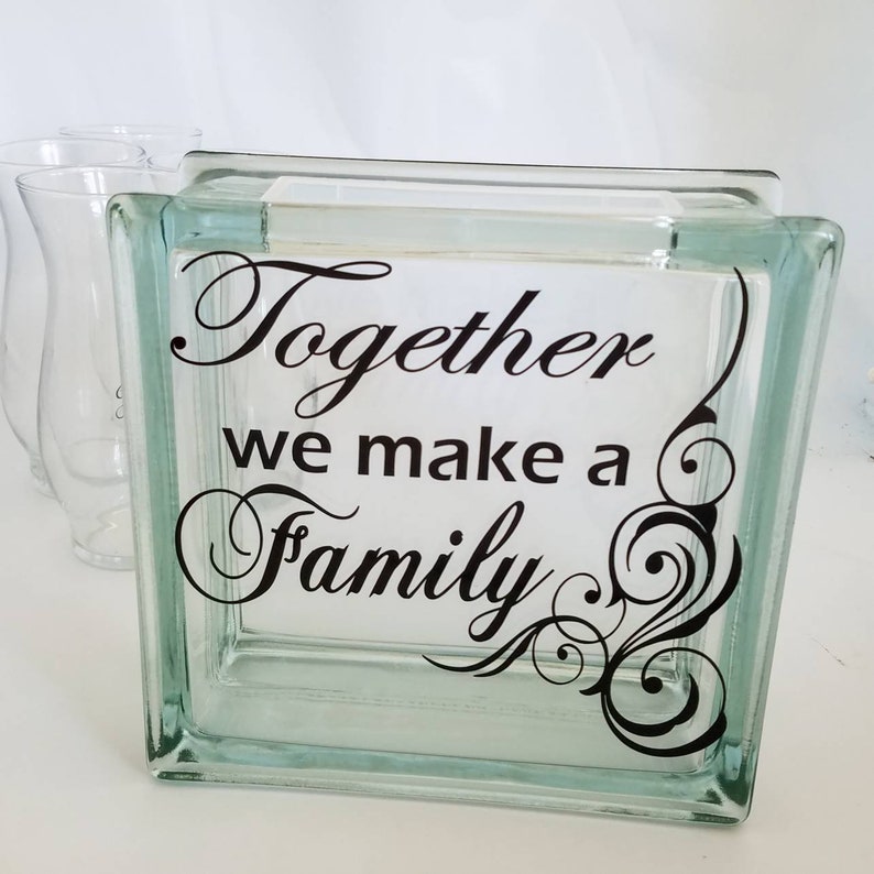 May include: A square, clear glass block featuring the black text Together we make a Family in a decorative script font. The design includes a black swirling flourish on the right side. Two clear glass vases are visible in the background.