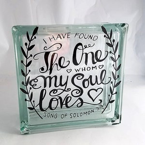 May include: A square, clear glass block featuring black lettering that reads, "I HAVE FOUND THE ONE WHOM MY SOUL LOVES" and "SONG OF SOLOMON 3:4". The text is framed by two vertical black leafy vine illustrations and small heart accents.