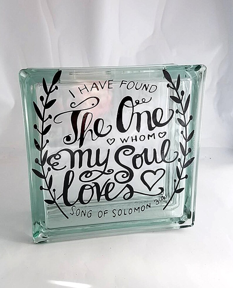 May include: A square, clear glass block featuring black lettering that reads, "I HAVE FOUND THE ONE WHOM MY SOUL LOVES" and "SONG OF SOLOMON 3:4". The text is framed by two vertical black leafy vine illustrations and small heart accents.