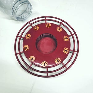May include: A red metal hummingbird feeder with a circular design and a clear glass jar. The feeder has a series of small, yellow flower accents around the center. The feeder is designed to attract hummingbirds.