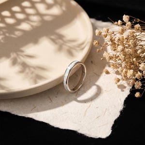 Op de afbeelding: Een zilverkleurige metalen ring met een centrale inleg van iriserend wit en lichtblauw materiaal dat aan opaal doet denken. De ring heeft een gladde, gepolijste afwerking en een afgerond profiel, met een subtiel kleurenspel in de inleg.