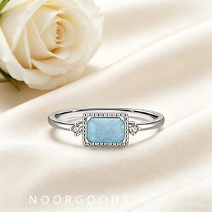 May include: A silver-toned ring featuring a rectangular, light blue opal-like stone set in a beaded frame. The band includes two small, clear, round stones on either side of the central setting. The text NOORGOODSCO is printed at the bottom.