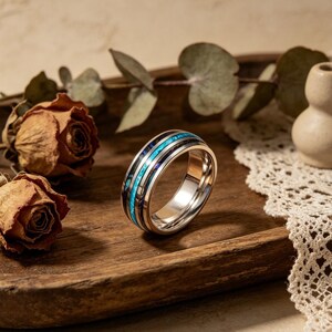 May include: A metallic band ring featuring a central inlay of iridescent blue and green abalone shell flanked by two thin black stripes. The ring has a polished silver-toned finish and a smooth, rounded profile, showcasing a reflective surface.