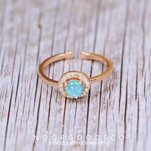 May include: Rose gold adjustable ring featuring a round, light blue opal center stone surrounded by a halo of small, clear crystals. The ring is open at the bottom and sits on a wooden surface. The text "NOORGOODSCO Handcrafted Jewellery" is at the bottom.
