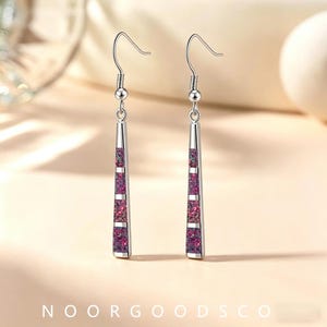 May include: A pair of silver-toned drop earrings featuring a long, slender, tapered metal frame. Each earring contains three rectangular inlays of iridescent, crushed purple and pink material, suspended from a simple curved ear wire hook.