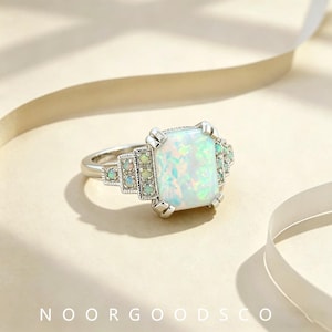 May include: A silver-toned ring featuring a large, rectangular, iridescent white opal center stone. The band is accented with smaller, round, iridescent opal stones set in a tiered, geometric pattern on both sides. The text NOORGOODSCO is visible.