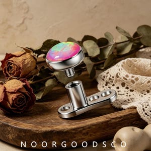 May include: A silver-toned dermal anchor body jewelry piece featuring a detachable top with a round, iridescent pink and green opal-like stone. The base is a flat metal plate with three circular holes, and the text NOORGOODSCO is visible.