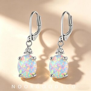 May include: A pair of silver-toned leverback earrings featuring oval-shaped iridescent opals with multicolored flashes. Each earring is accented with a single clear round-cut stone set above the opal, secured by four metal prongs.