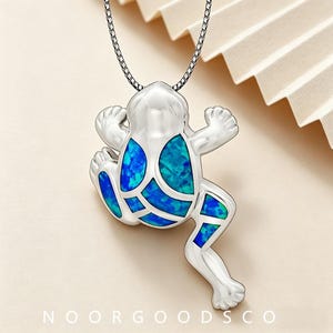 May include: A silver-toned frog pendant necklace featuring inlaid blue and green iridescent stone segments on the body and legs. The frog is suspended from a thin silver-toned chain. The text NOORGOODSCO is printed at the bottom of the image.