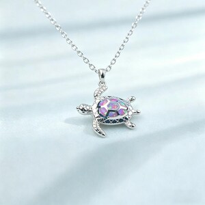 May include: A silver-toned necklace featuring a turtle pendant. The turtle shell is inlaid with a multi-colored iridescent opal stone that displays pink, blue, and purple hues. The pendant hangs from a delicate silver-toned chain link necklace.