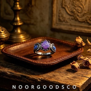 May include: A silver-toned ring featuring a large, round, purple cabochon stone at the center, flanked by two smaller, oval-shaped blue stones. The metal band has intricate detailing around the settings. The text NOORGOODSCO is visible.