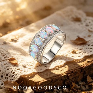 May include: A silver-toned ring featuring a row of five oval-shaped iridescent opals set vertically. The band is accented with small, clear pavé-set stones along the edges, creating a wide, polished metal design with a reflective finish.
