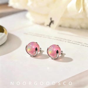 May include: A pair of stud earrings featuring spherical, translucent pink stones with iridescent highlights. Each stone is held by a silver-toned metal prong setting that curves around the base of the sphere. The earrings are displayed on a white surface.