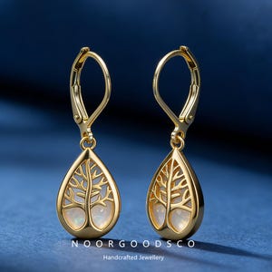 May include: Gold-tone teardrop earrings with a tree of life design. The earrings feature a lever back closure and a pearlescent center. The text "NOORGOODSCO" and "Handcrafted Jewellery" are visible.