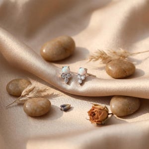 May include: A pair of silver stud earrings, each featuring a round white opal cabochon set above a cluster of three small, clear, round-cut stones. The earrings have a post-back closure and are displayed on a soft, draped, champagne-colored fabric.