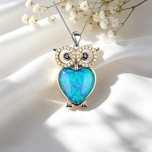 Könnte beinhalten: Silberne Eulen-Anhänger-Halskette mit herzförmigem, blauem Opal-Körper. Die Augen der Eule sind schwarz, und die Flügel sind mit kleinen, klaren Kristallen verziert. Der Anhänger hängt an einer zarten Silberkette.