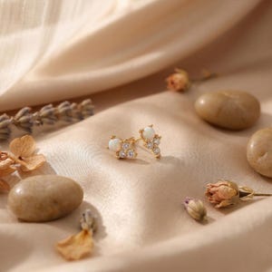 May include: A pair of gold stud earrings, each featuring a central round white opal cabochon accented by a cluster of three small, clear, round-cut stones arranged in a triangular formation, set in a polished gold metal base.
