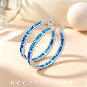 May include: A pair of silver-toned hoop earrings featuring a series of rectangular blue opal inlays set along the outer surface. The earrings have a hinged post closure mechanism and are positioned to show their circular shape and vibrant blue color.
