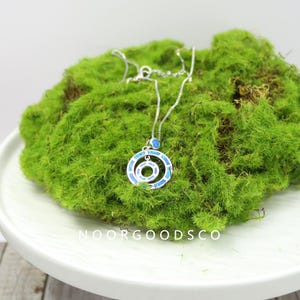 May include: A silver necklace with a pendant featuring concentric circles inlaid with blue opal-like stones. The necklace rests on a bed of vibrant green moss, with the text "NOORGOODSCO" visible on the white surface below.
