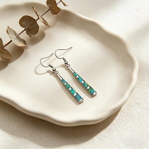 May include: A pair of silver-toned drop earrings featuring a long, slender bar design. Each earring is inlaid with four rectangular, iridescent blue-green stone segments. The earrings hang from standard curved silver-toned ear wires.