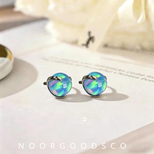 May include: A pair of stud earrings featuring spherical, iridescent blue opal-like stones held by dark metallic, curved prongs. The stones display vibrant flashes of green and pink light, set against a light surface with the text NOORGOODSCO.