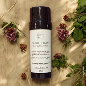 Puede incluir: Un recipiente cilíndrico marrón oscuro y blanco de loción Muscle Recovery, con el texto "Soothes Muscle Pain". La etiqueta también enumera los ingredientes y las instrucciones. El producto es de 71g. El fondo presenta flores secas y vegetación.