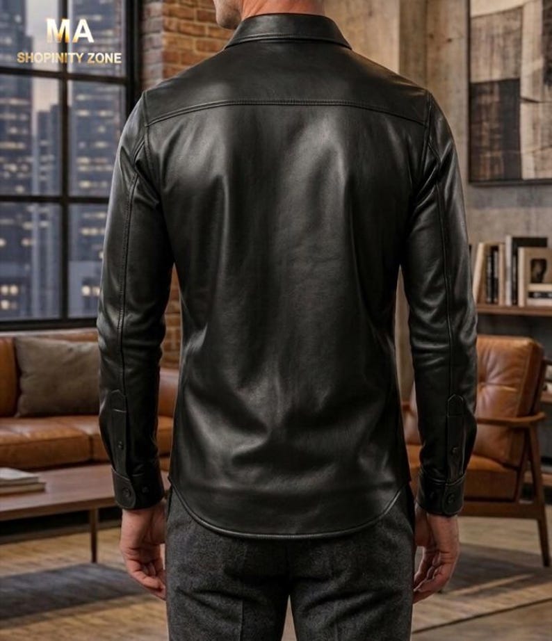 May include: A black leather shirt with long sleeves and buttoned cuffs. The shirt has a classic collar and a curved hemline. The back of the shirt is visible, showcasing the smooth texture of the leather. The shirt is worn with gray pants.