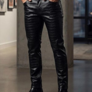 May include: Black trousers featuring a textured crocodile skin pattern, a button and zip fly closure, and front pockets. The trousers have a slim-fit silhouette and are paired with black leather boots. The text MA SHOPINITY ZONE is visible.