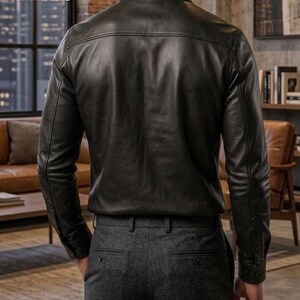 May include: A black leather shirt with long sleeves and buttoned cuffs. The shirt has a classic collar and a tailored fit. The model is wearing gray dress pants. The image is taken from the back, showcasing the shirt's design and texture.