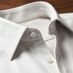 May include: Close-up of a white leather shirt with a classic collar and button-down front. The shirt features visible stitching and two white buttons. The texture of the leather is clearly visible, highlighting the garment's quality and design.
