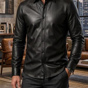 May include: Black leather button-down shirt with long sleeves. The shirt has a classic collar and is buttoned up the front. The shirt is worn with dark gray pants. The image includes the text "MA SHOPINITY ZONE".
