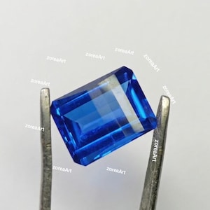 May include: A vibrant, rectangular, faceted blue gemstone held by silver tweezers. The gem has a deep, saturated color and a polished surface, reflecting light. The image includes the text "zoreaArt".