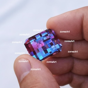 May include: A multi-coloured, rectangular-cut gemstone, showcasing a mosaic of blues, purples, and pinks. The gem is held by fingers, with the text "zoreaArt" visible on the gem's surface.