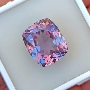 May include: A cushion-cut gemstone displaying a mix of pink, purple, and blue tones. The gem is presented against a white backdrop within a square container, emphasizing its detailed facets and colour variations.