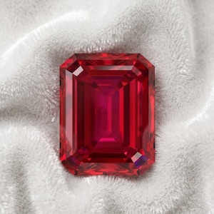 May include: A large, rectangular-cut ruby gemstone with a deep red hue. The gem is set against a soft, white, textured background, highlighting its facets and brilliance. The ruby's color is consistent throughout, with a slightly darker center.