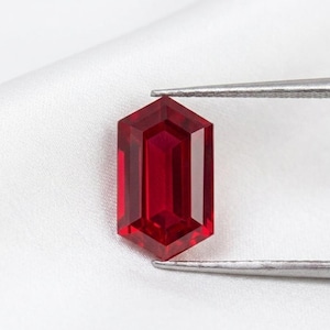 May include: A faceted, deep red gemstone with a hexagonal shape. The gem is held by silver tweezers against a white background. The gem's facets reflect light, showcasing its clarity and color.
