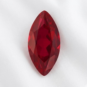 May include: A faceted, marquise-cut gemstone in a deep, rich red hue. The gem's elongated shape and multiple facets create a brilliant display of light and shadow. The gemstone is set against a white background.