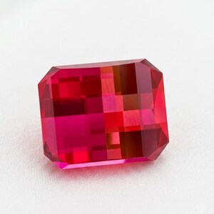 Puede incluir: Una piedra preciosa rectangular facetada de un color rojo intenso y vibrante. La piedra presenta un diseño de corte escalonado con bordes limpios y un acabado transparente de alto brillo que refleja la luz a través de su estructura geométrica.