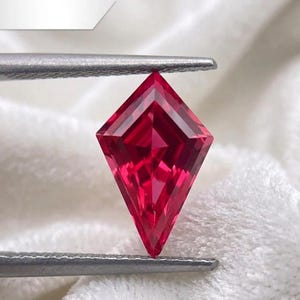 May include: A faceted, kite-shaped gemstone in a vibrant red hue. The gem is held by silver tweezers against a soft, white background. The cut of the stone creates multiple facets, reflecting light and enhancing its brilliance. This gemstone is likely for jewelry making.