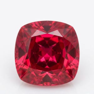 May include: A cushion-cut gemstone with a vibrant, deep red color. The stone features a faceted surface that creates a complex geometric pattern of light and dark reflections across its square shape with rounded corners and a polished finish.