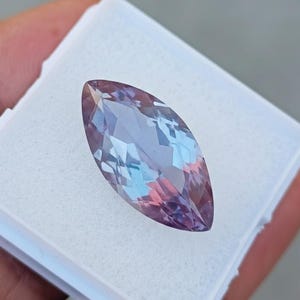 May include: A marquise-cut gemstone, displaying a blend of purple, blue, and pink colours. The gemstone is faceted, creating multiple reflective surfaces. It is set in a white plastic container, held by a hand.
