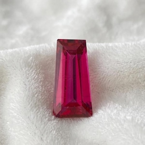May include: A faceted, rectangular gemstone with a deep magenta hue. The gem has a trapezoidal shape with a flat top and a series of internal facets that create a play of light and shadow. The gemstone is set against a white, textured background.