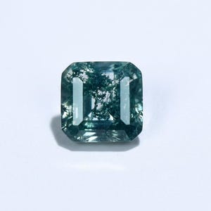 May include: A square-cut, moss agate gemstone with a translucent, teal-green hue. The stone features internal inclusions resembling a miniature landscape. The gem is set against a white background, highlighting its clarity and color.