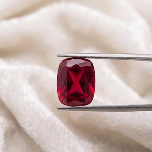 May include: A vibrant, cushion-cut ruby gemstone, held by silver tweezers against a soft, white background. The ruby displays a deep red hue with internal facets reflecting light. The gemstone's shape is rectangular with rounded corners.