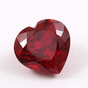 Peut inclure: Une pierre précieuse en forme de cœur, taillée à facettes, d'un rouge profond et riche. La gemme présente de multiples facettes qui captent la lumière, créant un effet scintillant. La forme est symétrique, avec une pointe claire en bas et une courbe douce en haut.